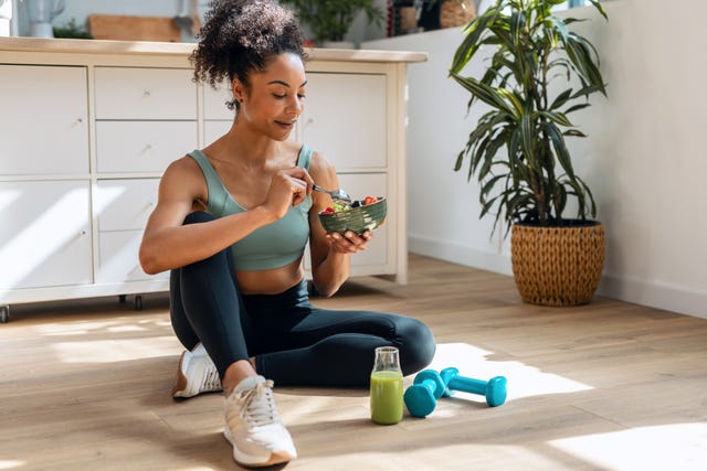 shot of athletic woman eating a healthy fruit bowl while sitting on floor in the kitchen at home