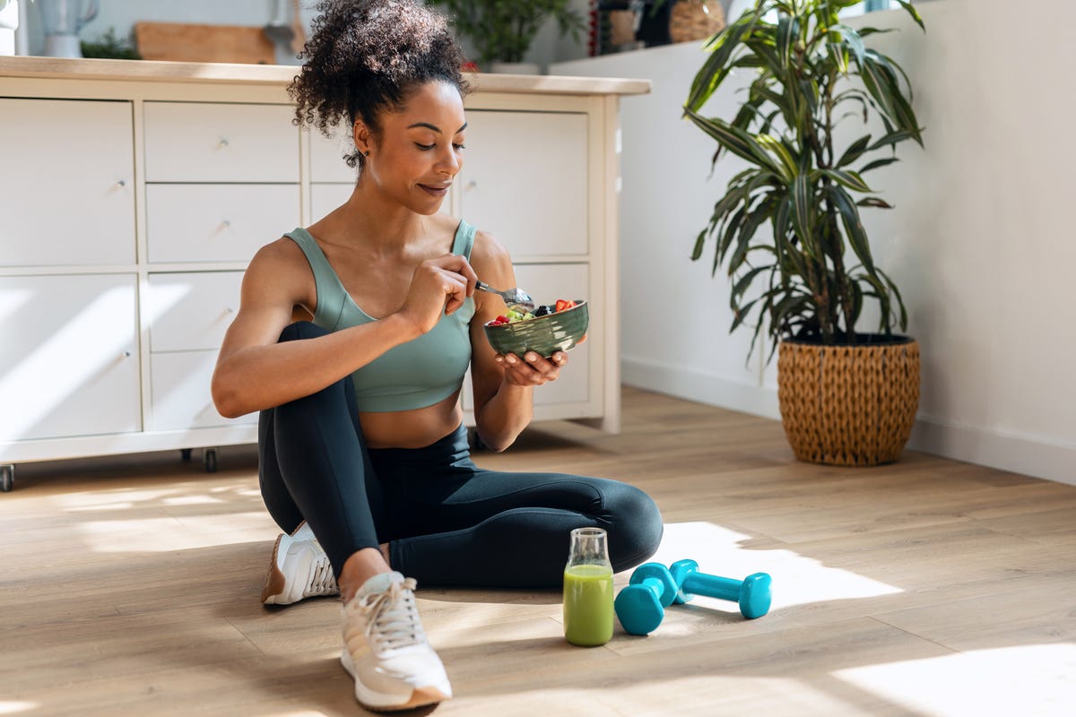 shot of athletic woman eating a healthy fruit bowl while sitting on floor in the kitchen at home