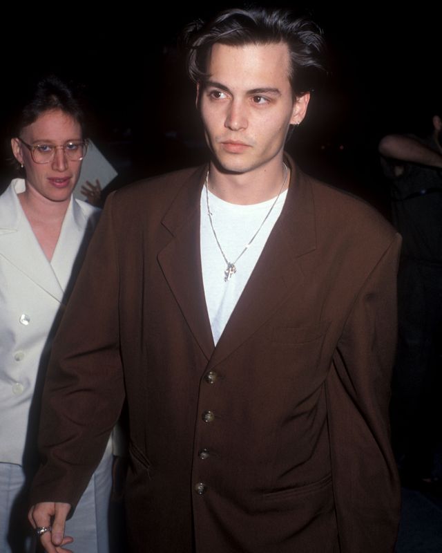 johnny depp walks and looks past the camera, he wears a white tee shirt, brown baggy suit jacket and silver cross necklace