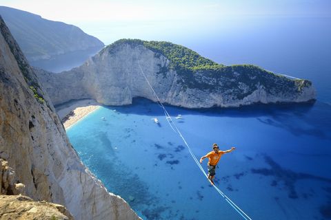 Een slackliner loopt over een touw over het strand van Navagio in Zakynthos Griekenland Slacklining is een sport waarbij een plat touw wordt gespannen tussen twee punten vaak met minder spanning dan een koord Degenen die overheen lopen dragen harnassen verbonden met de slackline om ze op te vangen als ze vallen