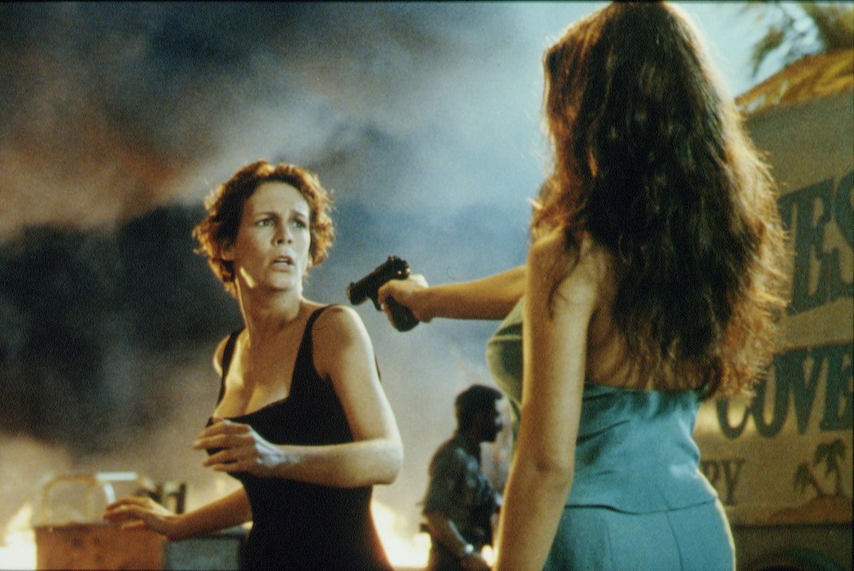 a publicity photo from the movie true lies, featuring jamie lee curtis reacting with concern as a woman points a gun at her