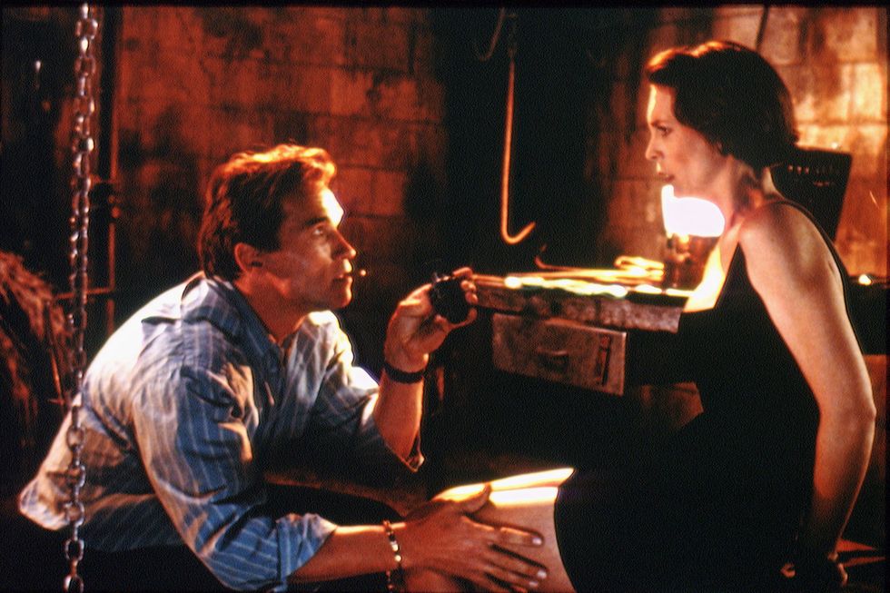 arnold schwarzenegger and jamie lee curtis appear in a scene of the film true lies, a kneeling schwarzenegger is looking up at a frightened curtis, he touches her thigh and holds an unidentified object in his other hand, curtis has her hands tied behind her back, they are in workshop looking area