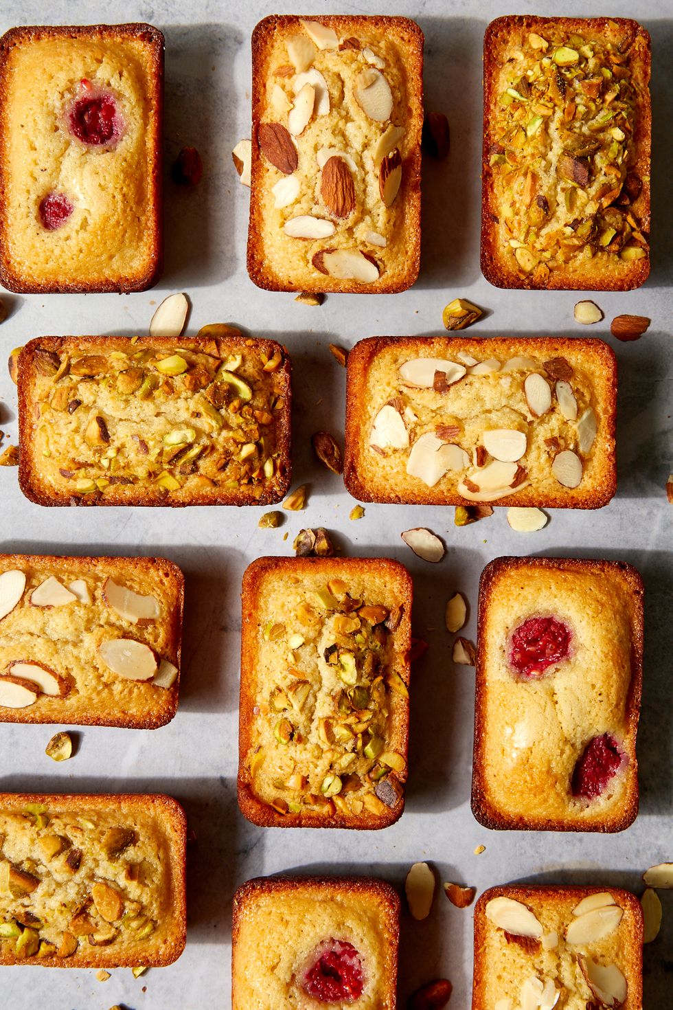 a group of financiers topped with different ingredients like raspberries, pistachios, and sliced almonds