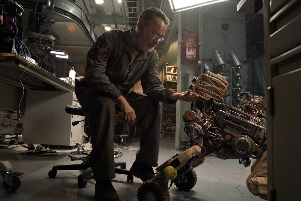 A person crouches and reaches for a small wheeled robot equipped with mechanical components and a device in his hand