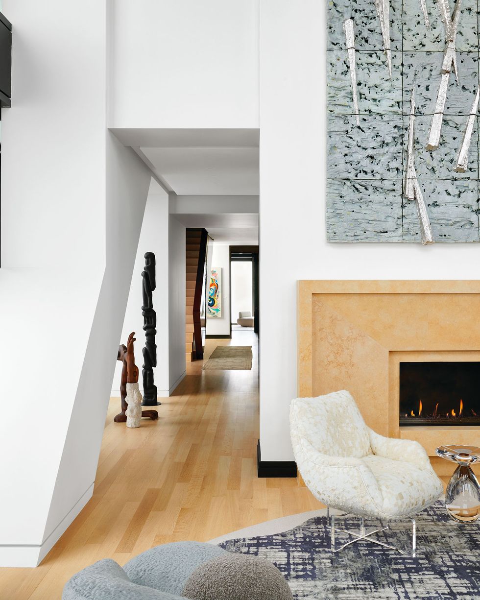 the living room with a horizontal fireplace and 3d artwork hanging above it, a patterned rug and light colored club chair with an acrylic side table, a black console by the window and totems in a hallway