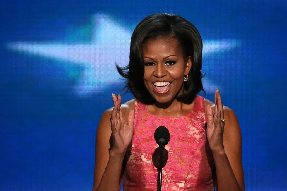 michelle obama, wearing a pink dress, speaking into a camera, with a blue and white backdrop behind her