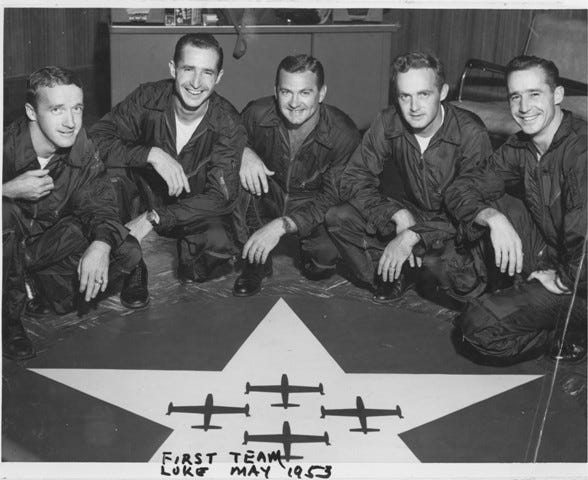 group of five individuals in flight suits posing on a starpatterned surface with representations of aircraft