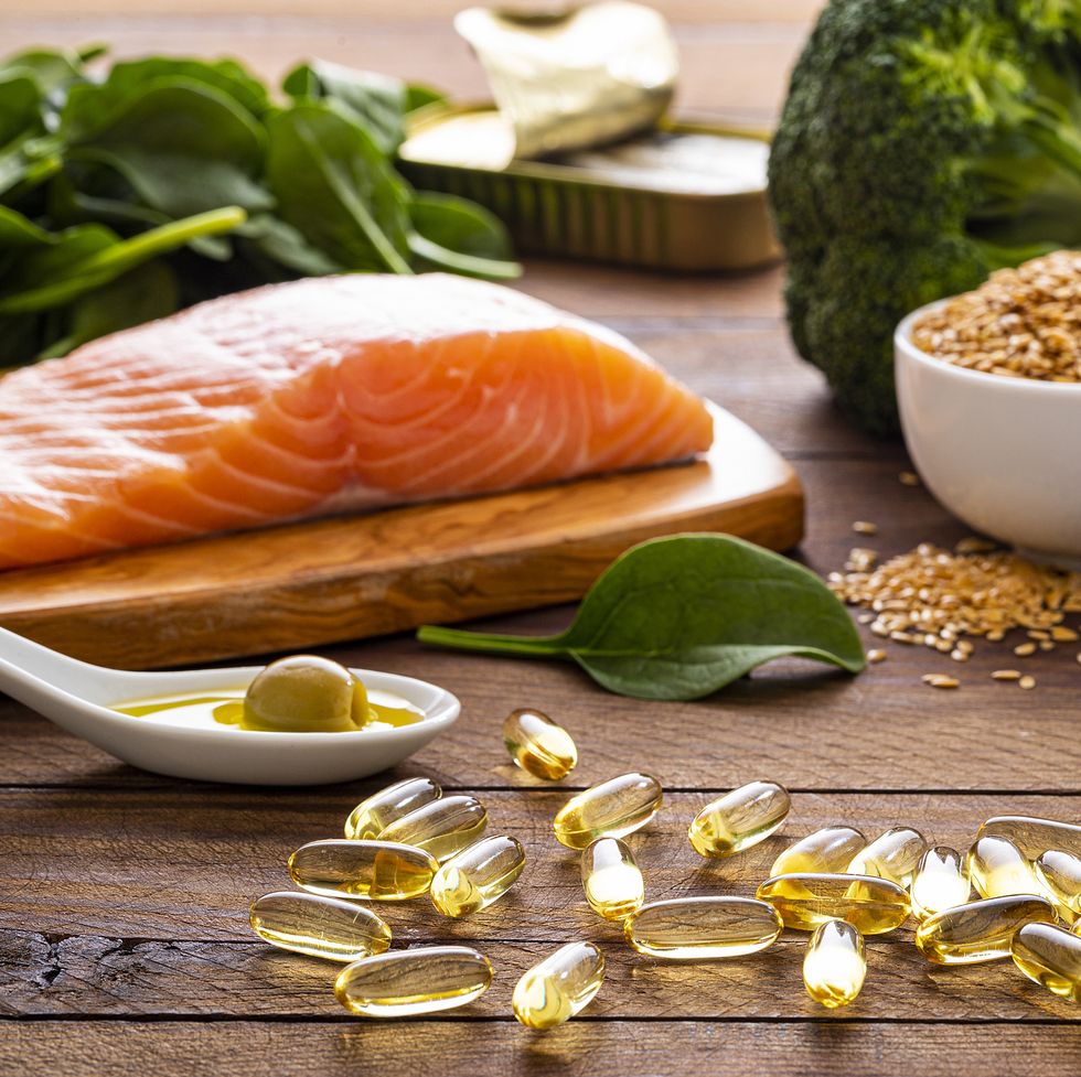 front view of many fish oil capsules spilling out from the bottle surrounded by an assortment of food rich in omega 3 such as salmon, flax seeds, broccoli, sardines, spinach, olives and olive oil