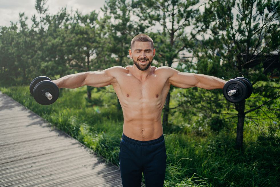 fit adult man lifts heavy weight during workout session outdoor, poses with naked torso, has well shaped muscular body, demonstrates his power, lifts barbells motivated bodybuilder at street
