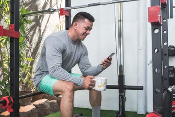 a fit asian man browsing his social media or chatting while seated on the squat rack holding a mug of coffee at a home gym