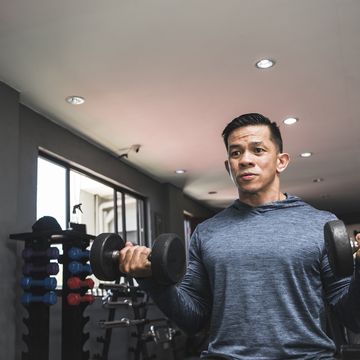 a fit asian man in a sweatshirt does seated dumbbell curls working out and training biceps open air gym setting