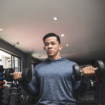 a fit asian man in a sweatshirt does seated dumbbell curls working out and training biceps open air gym setting