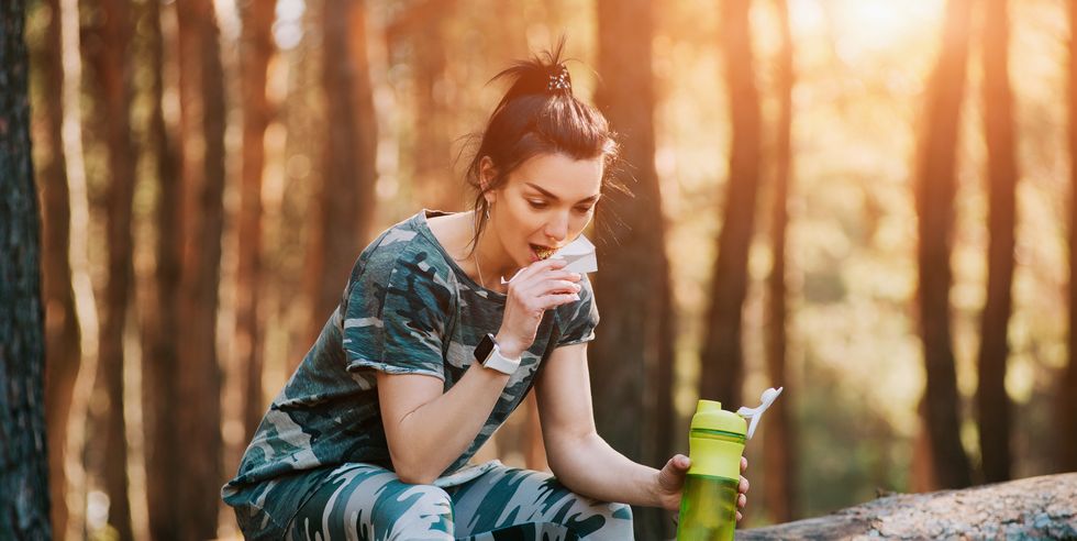 fit woman eating energy granola bar after running at sunset in the forest