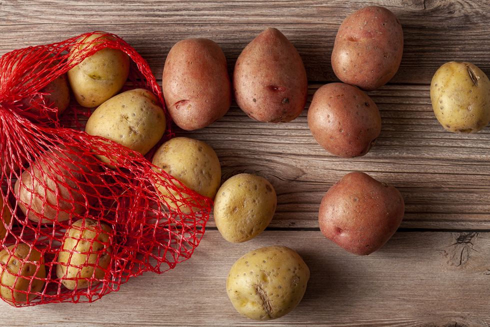 flat lay close up image featuring a red mesh potato sack with pink and yellow raw organic potatoes on wooden background