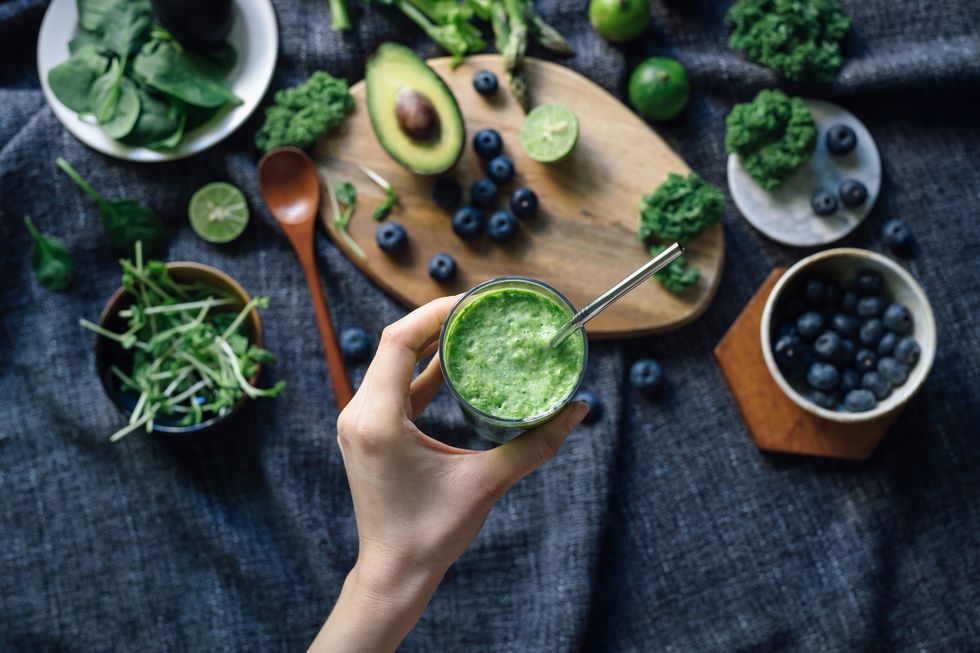flat lay of fresh and healthy green fruits and vegetables on a rustic wooden board, against dark grey fabric for a healthy eating dietwoman hand holding a glass of green smoothie with reusable straw healthy superfood green colour detox diet concept