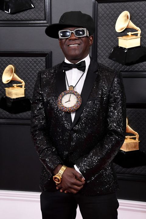 flavor flav wears a sequined black blazer, black pants, white shirt, black bow tie, and jeweled clock necklace on the red carpet, along with jeweled sunglasses and a black baseball cap