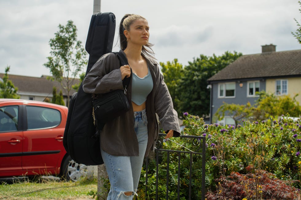 A person stands on a path carrying a guitar case over his shoulder and a bag in his hand