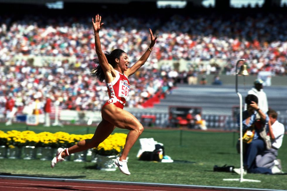 florence joyner runs on a track with her arms straight over her head, she smiles and wears a red and white singlet, white shoes and a number on her chest, yellow flowers and crowded stands are in the background