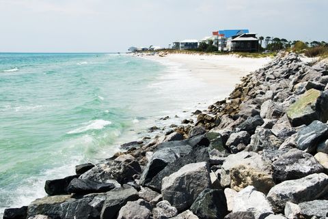 boulders in foreground leading to white sand beach and blue water with people and houses beyond