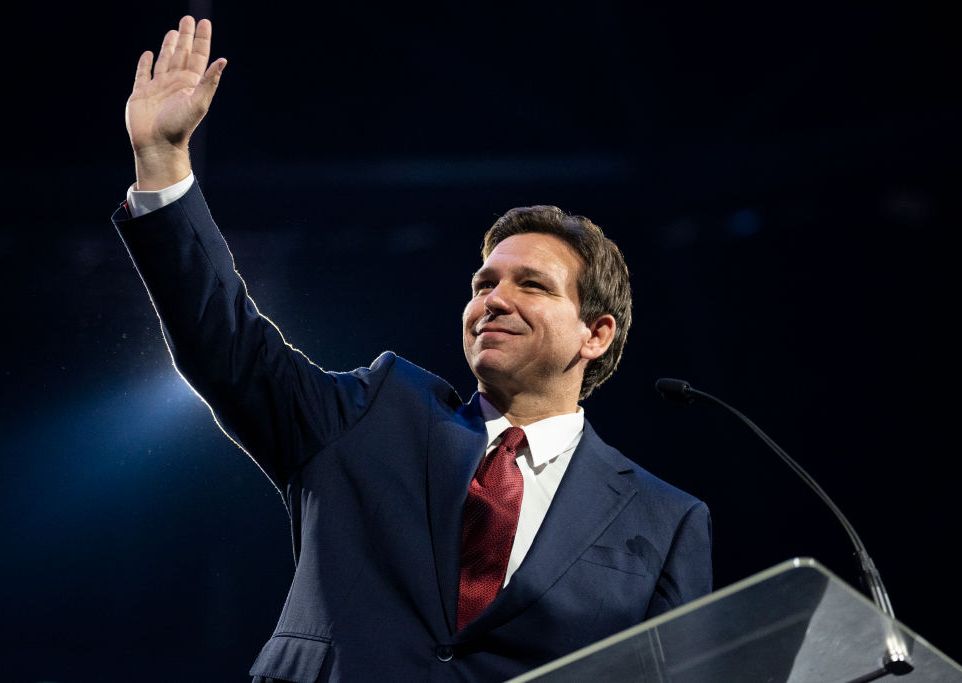 ron desantis waving to an audience with his right hand as he stands at a podium