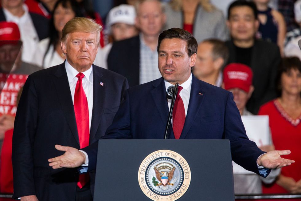 donald trump stands on a stage behind ron desantis who speaks at a lecturn, both men wear suits with white collared shirts and red ties