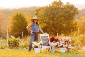niki irving standing next to buckets of dahlias on her farm, wearing cropped jeans, flutter sleeve blue shirt, brimmed sunhat