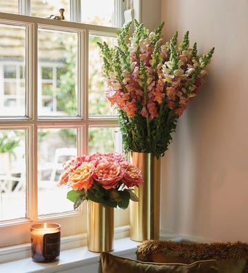 two flower arrangements in gold vases near a window next to a candle in a roundup of the best flower subscriptions 2024