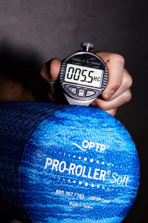 a hand is holding a silver durometer that displays a hardness measurement of 555 on its digital screen above a blue foam roller