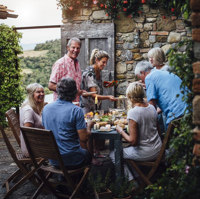 a group of mature friends are sitting around an outdoor dining table, eating and drinking they are all talking happily and enjoying each others company the image has been taken in tuscany, italy