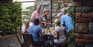 a group of mature friends are sitting around an outdoor dining table, eating and drinking they are all talking happily and enjoying each others company the image has been taken in tuscany, italy