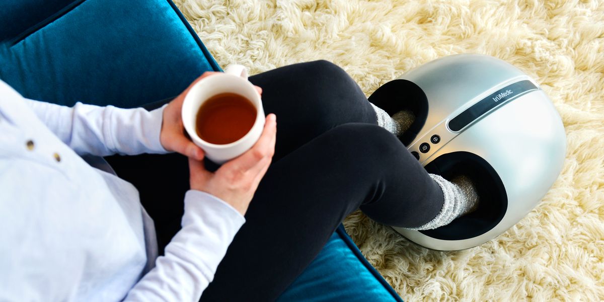 person sitting on couch holding a cup of tea with feet in a foot massager