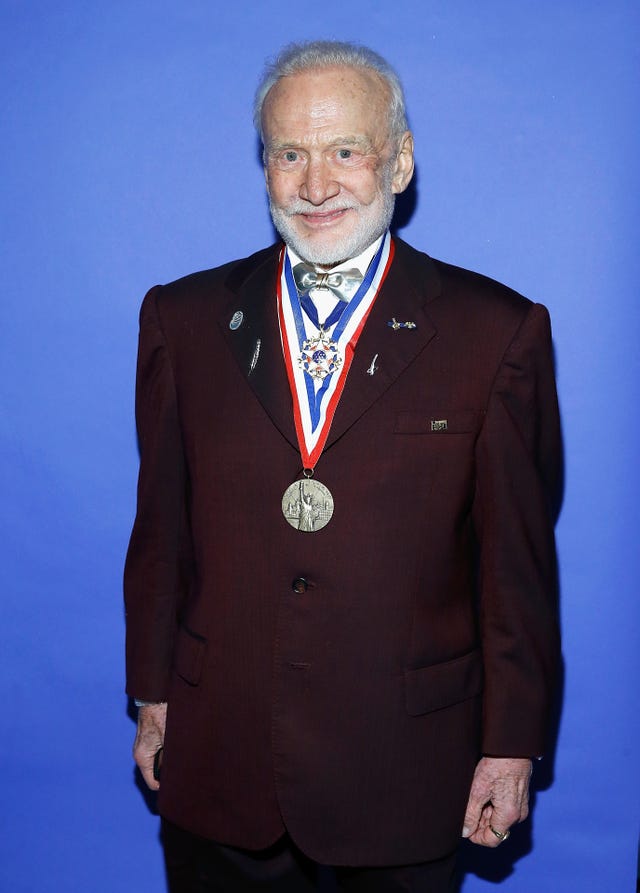 buzz aldrin smiles at the camera, he wears a maroon suit jacket, white collared shirt and silver bowtie with several medals hanging around his neck