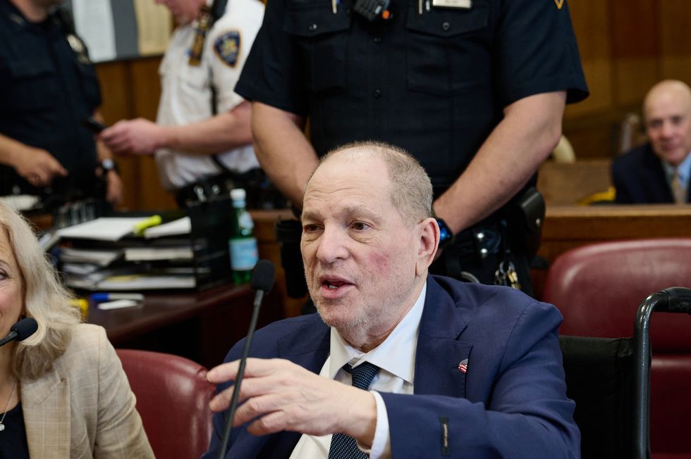 a man in a blue suit jacket and tie with a white collared shirt sits in a wheelchair at a wooden table and speaks while pointing one hand forward