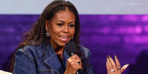 michelle obama speaks into a microphone while sitting in an arm chair, she wears a denim outfit, earrings and three rings, a book with sticky notes sits next to her on the chair