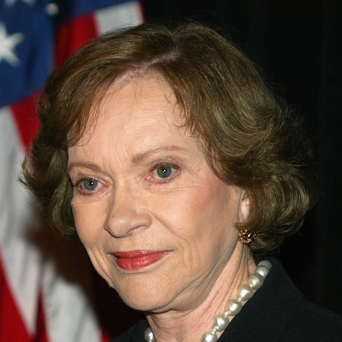 rosalynn carter stares past the camera with a slight smile on her face, she is wearing eye makeup, blush, and soft red lipstick, her brown hair is styled short with small curls away from her face, part of her large pearl necklace is visible, as is one of her gold earrings, in the blurred background is a black backdrop and an american flag