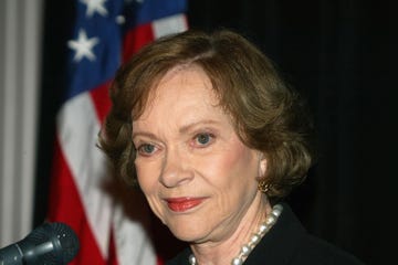 rosalynn carter stares past the camera with a slight smile on her face, she is wearing eye makeup, blush, and soft red lipstick, her brown hair is styled short with small curls away from her face, part of her large pearl necklace is visible, as is one of her gold earrings, in the blurred background is a black backdrop and an american flag