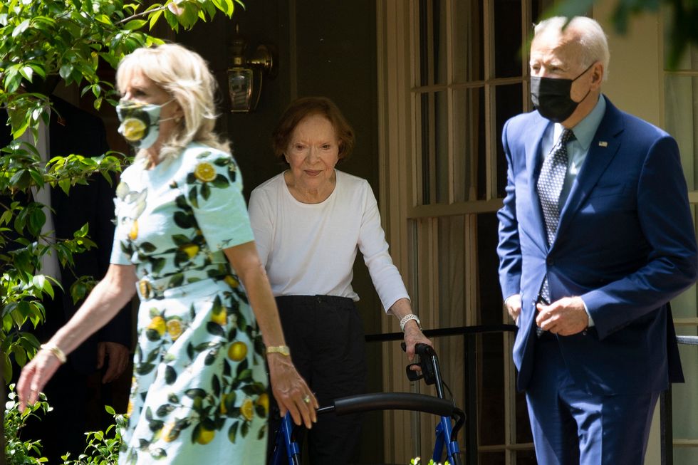 rosalynn carter uses a walker as she moves next to a window, two people walk in the forefront of the photo frame