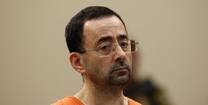 larry nassar stands and looks below the camera, he wears an orange inmate shirt, white undershirt and rectangular glasses