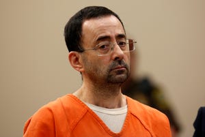 larry nassar stands and looks below the camera, he wears an orange inmate shirt, white undershirt and rectangular glasses