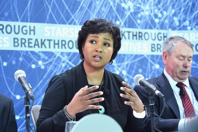 mae jemison speaks into a microphone as she sits at a table with a person on either side, she looks to the left and has her hands in front of her chest, she wears a black outfit with jewelry