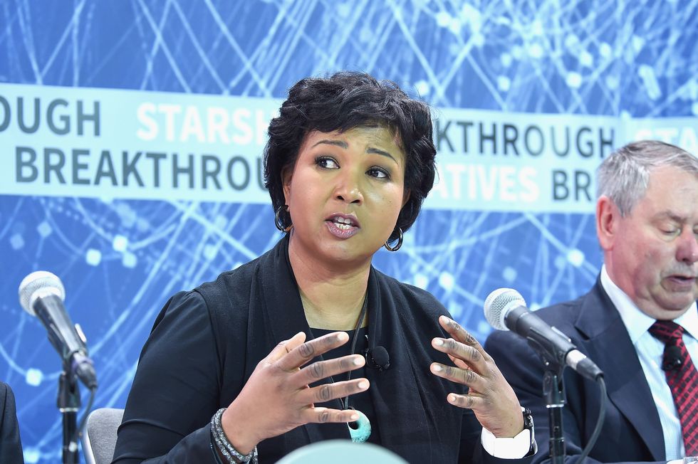mae jemison speaks into a microphone as she sits at a table with a person on either side, she looks to the left and has her hands in front of her chest, she wears a black outfit with jewelry