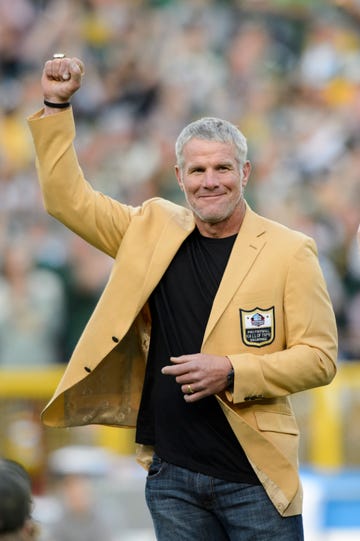 brett favre smiles and looks right as he raises one fist in the air, he wears a yellow suit jacket with a pro football hall of fame patch on the pocket and a black shirt