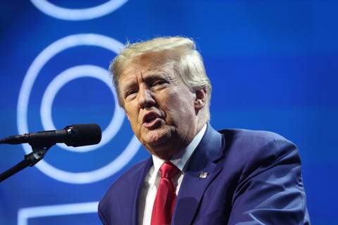 donald trump, wearing a blue suit jacket and red tie, speaking into a microphone with a blue backdrop behind him