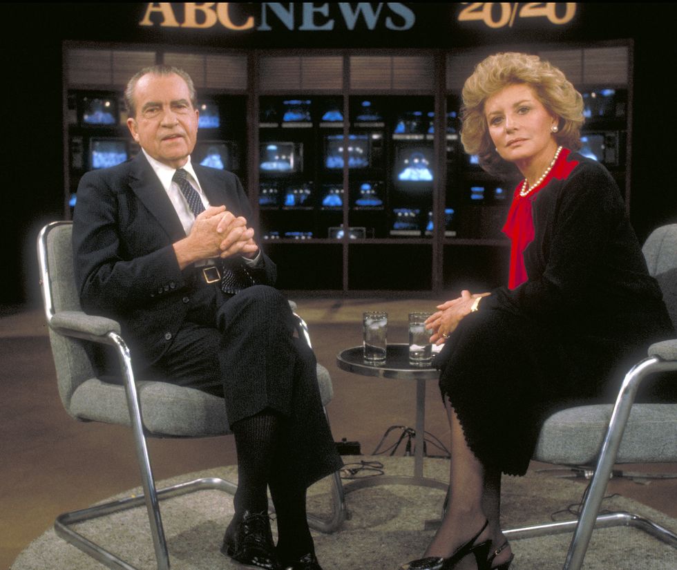 richard nixon and barbara walters sit in grey office chairs on a tv set and look at the camera, both are wearing dark suits but walters has on a bright red shirt, behind them is a side table with two water glasses, a wall of tv monitors, and a sign that says abc news 20 20