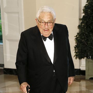 henry kissinger smiles at the camera, he wears a black suit with a black bowtie and a white collared shirt, he holds onto a cane while standing in a room