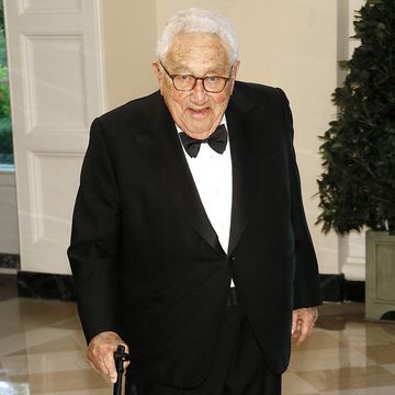 henry kissinger smiles at the camera, he wears a black suit with a black bowtie and a white collared shirt, he holds onto a cane while standing in a room
