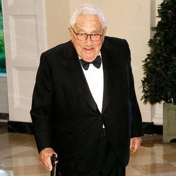 henry kissinger smiles at the camera, he wears a black suit with a black bowtie and a white collared shirt, he holds onto a cane while standing in a room