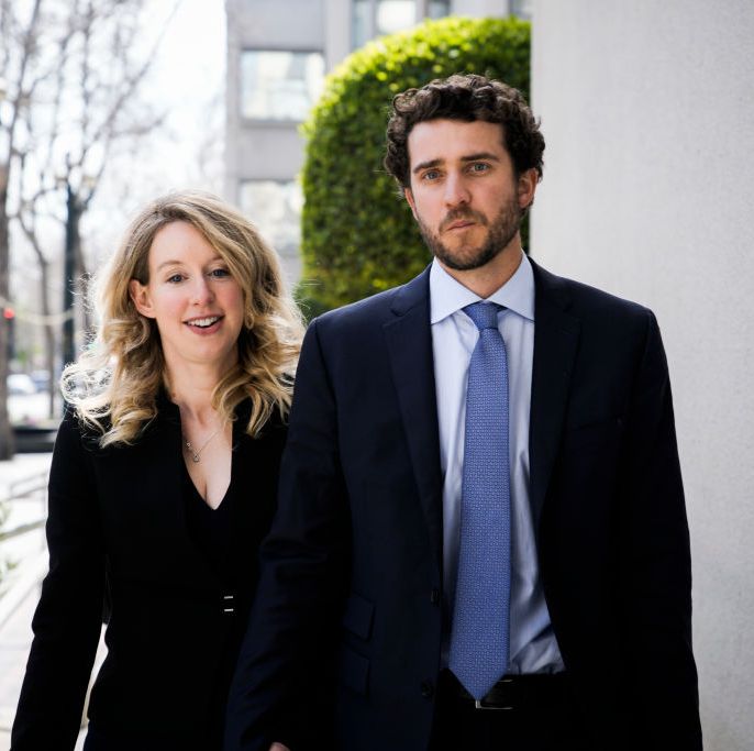 elizabeth holmes and billy evans walk next to each other outside a gray building, she is wearing an all black outfit and smiling, he is wearing a black suit jacket with blue shirt and tie and looks directly at the camera
