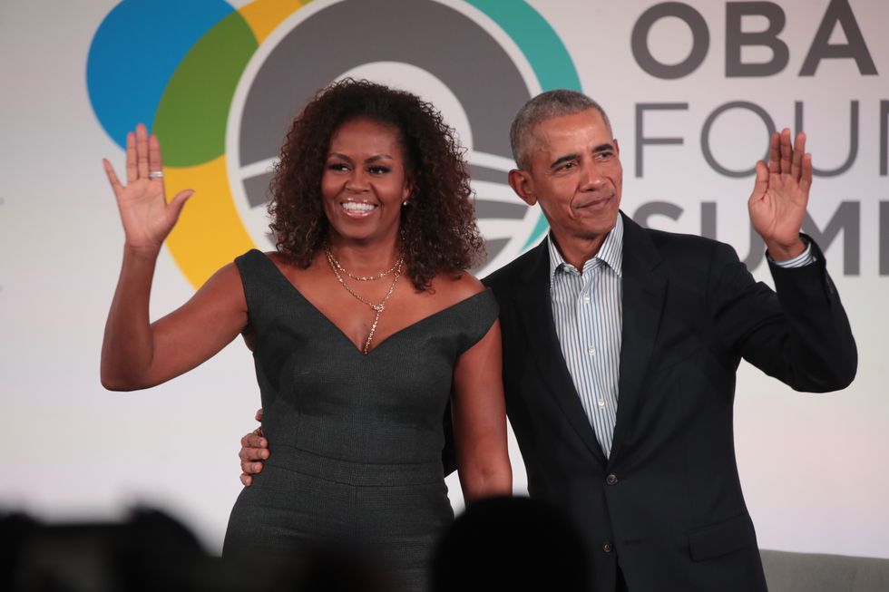 michelle and barack obama wave and smile to the off camera audience and stand in front of a colorful logo and the words obama foundation summit