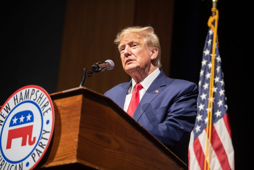 donald trump wearing a blue suit and red tie, speaking into a microphone at a podium, with an american flag behind him